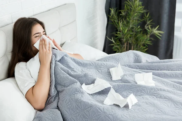 young sick woman sitting in bed bowing out her running nose with paper towel feeling bad early in the morning