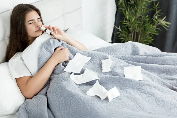 young sick woman laying in bed spraying medicine into her running nose using paper towels in the morning