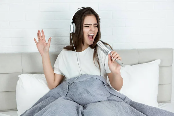 Joven hermosa mujer en auriculares cantando fuerte usando cepillo de pelo como un micrófono sentado en la cama por la mañana — Foto de Stock