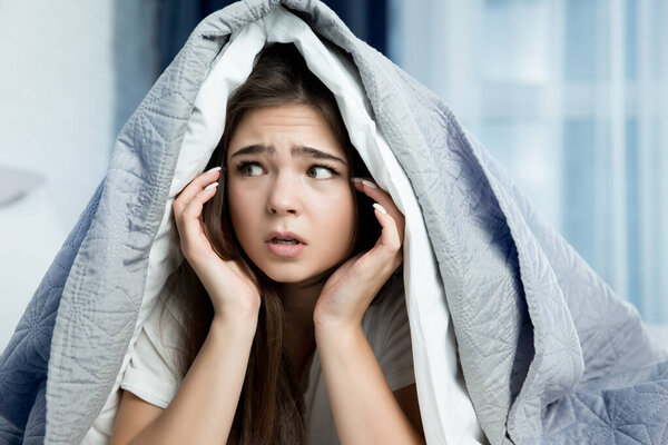 young beautiful brunette woman sitting in bed with blanket on her head looking scared in the morning