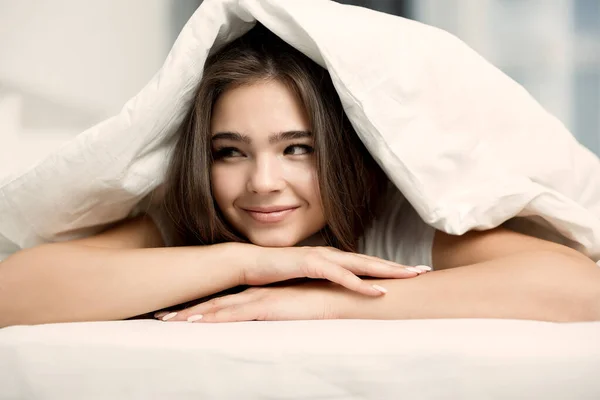 Wanita muda cantik berambut cokelat tersenyum duduk di tempat tidur dengan selimut di atas kepalanya tampak santai di pagi hari — Stok Foto