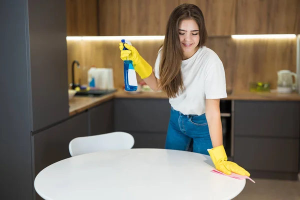 Joven hermosa mujer en guantes amarillos con aerosol detergente en la mano haciendo limpieza de limpiar el polvo de la mesa de la cocina con un trapo — Foto de Stock