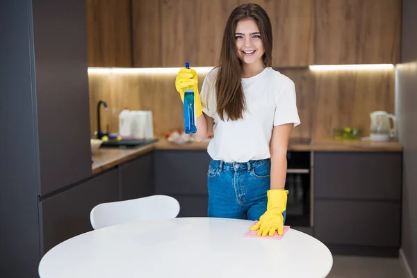 Joven hermosa mujer sonriente en guantes amarillos con spray detergente en la mano haciendo limpieza de limpiar el polvo de la mesa de la cocina con trapo — Foto de Stock