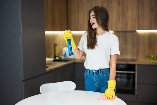 Schattig jong vrouw in geel handschoenen met wasmiddel spray in haar hand doen schoonmaken vegen stof af van de keukentafel met een doek — Stockfoto