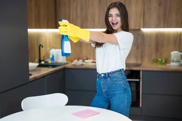 Linda joven con guantes amarillos con detergente rociado en broma mientras limpia el polvo de la mesa de la cocina con un trapo — Foto de Stock