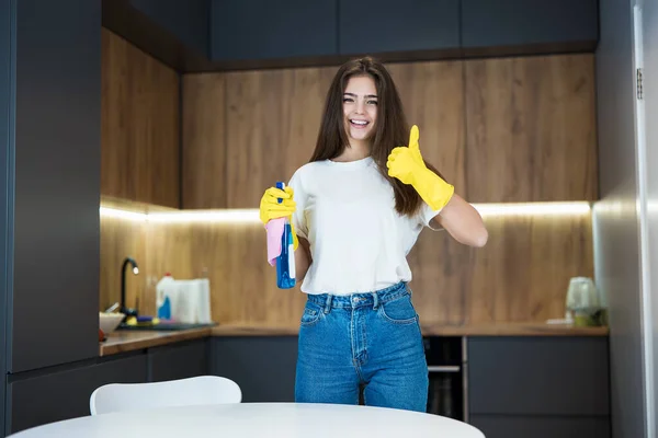 Linda joven con guantes amarillos con spray detergente en la mano limpiando el polvo de la mesa de la cocina con un trapo que muestra como signo — Foto de Stock