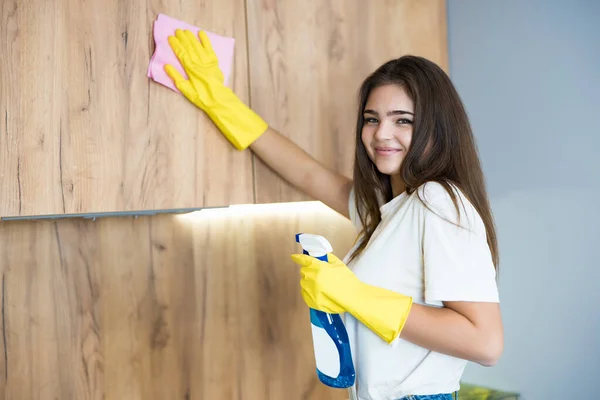 Hermosa joven sonriente con guantes amarillos con spray detergente en la mano limpiando el polvo del gabinete de la cocina con un trapo — Foto de Stock