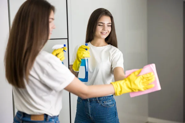 Beautiful young smiling woman in yellow gloves with detergent spray in her hand wiping mirror glass with a rag — Stock Photo, Image