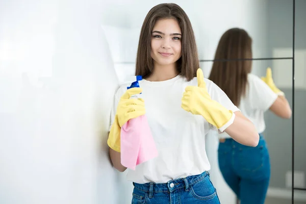 Cute young woman in yellow gloves with detergent spray in her hand and rag in another standing near mirror smiling showing like sign — Stock Photo, Image