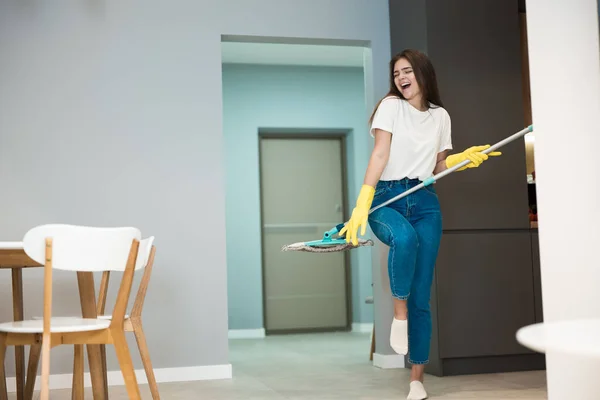 Забавная молодая женщина в желтых перчатках танцует со шваброй и поет во время мытья пола на кухне, чувствуя себя счастливой, притворяясь рок-звездой — стоковое фото