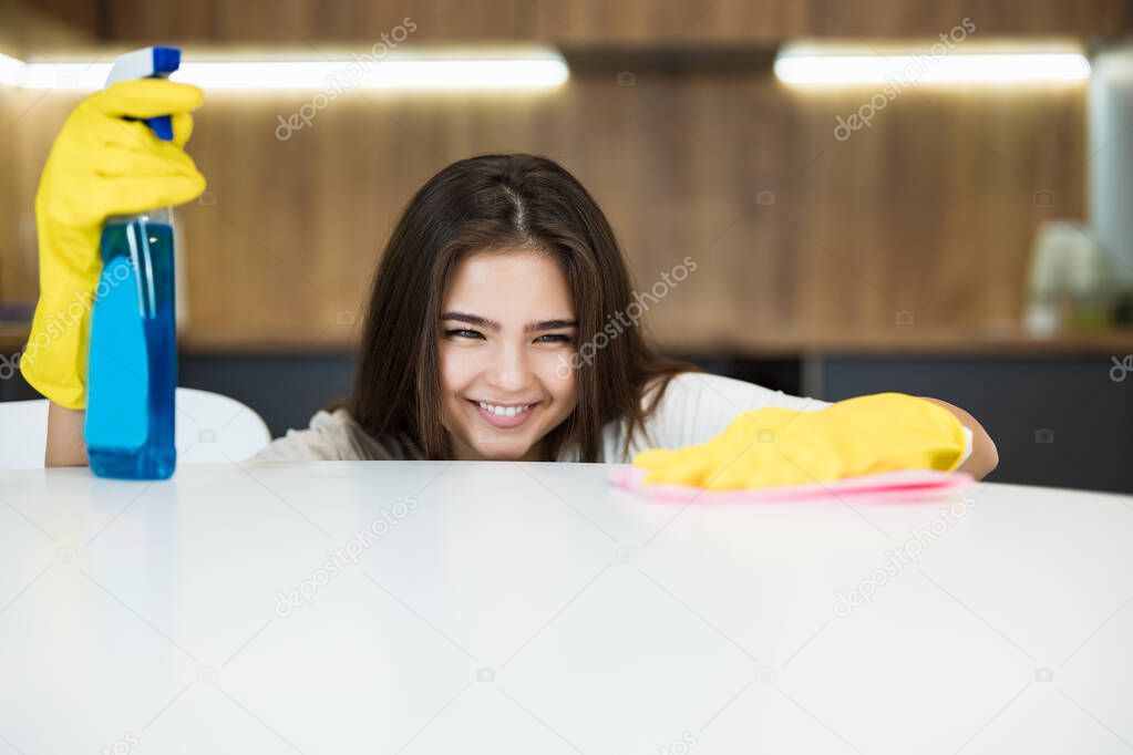 beautiful smiling young woman in yellow gloves with detergent spray in her hand thoroughly wiping dust off from the kitchen table with a rag