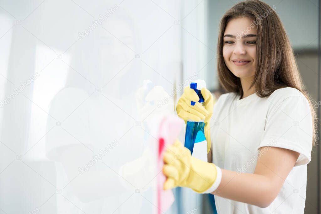 cute young woman in yellow gloves spraying detergent spray while wiping dust off from the kitchen wall with a rag thoroughly