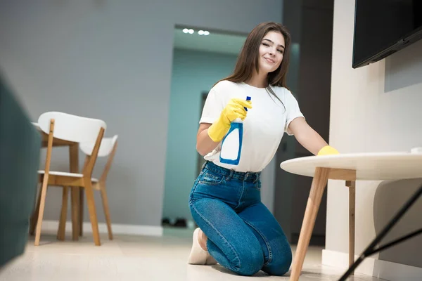 Cute young woman in yellow gloves with detergent spray in her hand wiping dust off from the kitchen chair with a rag — Stock Photo, Image