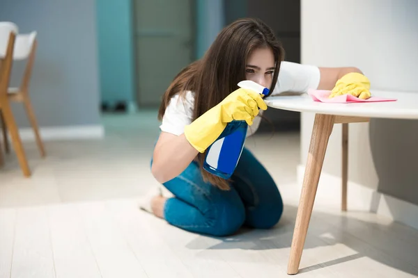 Cute young woman in yellow gloves with detergent spray in her hand wiping dust off stain from the kitchen chair with a rag thoroughly — Stock Photo, Image