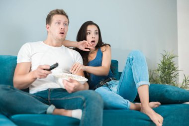 Genç çift güzel esmer kadın ve yakışıklı adam kanepede korku filmi izleyip patlamış mısır yiyorlar.