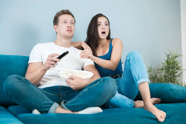 Jeune couple belle femme brune et bel homme regardant un film sur le canapé manger du maïs pop à la recherche d'un style de vie confortable surpris — Photo