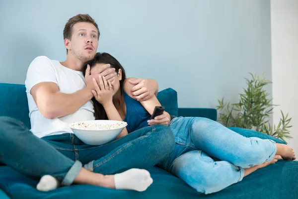 Young couple beautiful brunette woman and handsome man watching scary movie on the sofa eating pop corn looking frightened he closes her eyes homesters — Stock Photo, Image