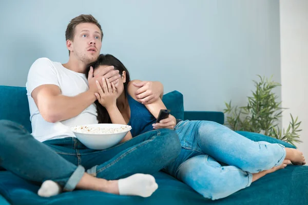 Jong paar mooi brunette vrouw en knappe man kijken eng film op de bank eten pop maïs kijken bang hij sluit haar ogen homesters — Stockfoto