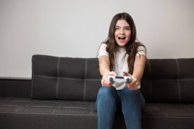 Kumral genç bir kadın elinde joystick ile kanepede otururken mutlu görünüyor. Oyun oynuyor, kendini evinde gibi hissediyor..