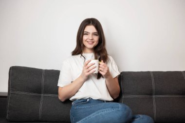 Genç, güzel, gülümseyen esmer kadın, elinde çay ve kahveyle kanepede oturuyor. Mutlu görünüyor, evinde rahat hissediyor..