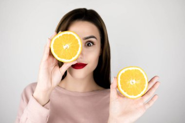 Kızıl dudaklı genç esmer kadın bir elinde turuncunun yarısını diğer elinde tutuyor, güzellik anlayışı, izole edilmiş beyaz arka plan.