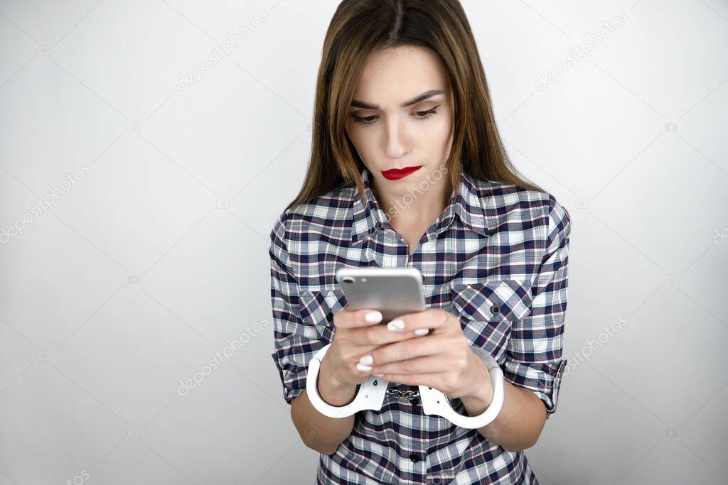 brunette young woman holds smartphone in her hands, texts while having handcuffs on , virtual reality concept, social media addiction, nomophobia, isolated white background.