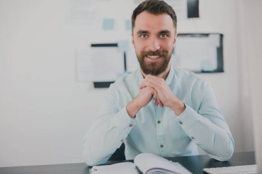 Genç, sakallı, gülümseyen esmer adam modern ofisinde iş projesinde çalışırken mutlu görünüyor, yüzünün yanında el ele tutuşuyor, rutin iş kavramı..