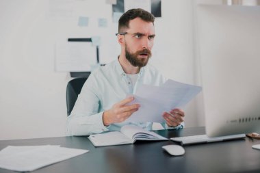 Genç sakallı esmer adam modern ofisinde iş projesinde çalışırken çok şaşırmış görünüyor elinde belgelerle, rutin iş kavramı.