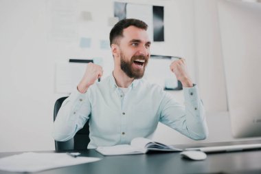 Genç sakallı esmer adam başarılı bir iş anlaşmasından sonra mutlu görünüyor, modern ofisinde elleri kazanan pozisyon, başarı ve refah konseptinde oturuyor..
