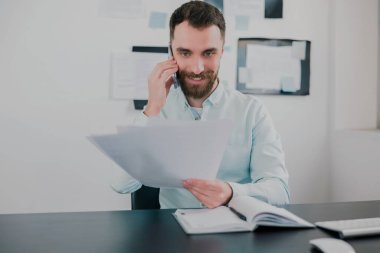 Genç sakallı esmer adam modern ofisinde iş projesinde çalışıyor elinde belgeler, telefonla konuşuyor, rutin iş kavramı..