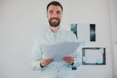 Genç, sakallı, esmer, gülümseyen bir adam modern ofisinde iş projesinde çalışıyor, iki elinde de belgeler var, rutin çalışma konsepti..