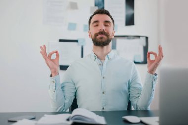 Modern ofisteki bilgisayarında iş projesi üzerinde çalışırken sakin görünen sakallı genç adam, huzurlu bir yoga pozisyonunda el ele tutuşuyor, işte rahatlıyor..