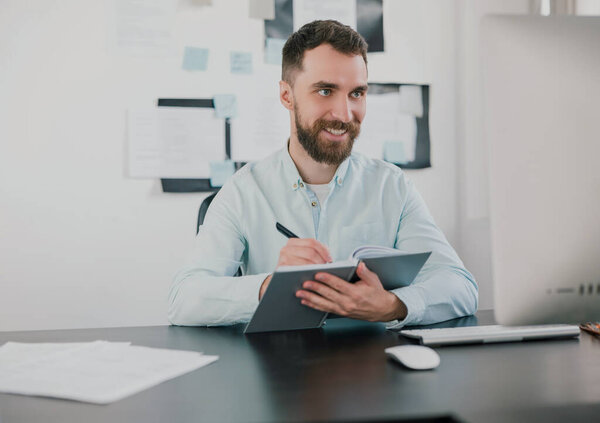 young bearded brunette smiling man looking happy while taking notes to his agenda working on business project in his modern office, work routine concept.