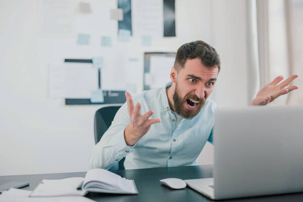 молодой бородатый брюнетка мужчина выглядит сердитым, работая над бизнес-проектом в своем ноутбуке в современном офисе, пожимая руки, рабочая рутинная концепция.