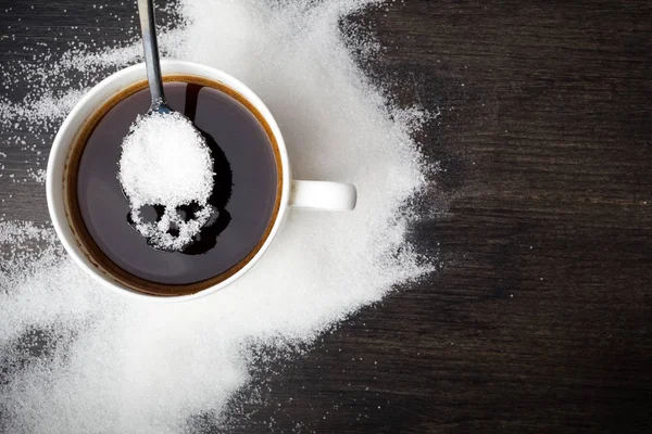 Unhealthy white sugar concept. Scull spoon with sugar and cup of black coffee on wooden background — Stock Photo, Image