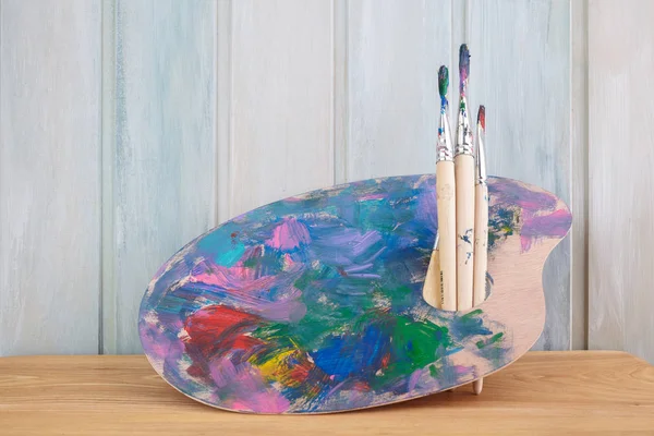 木制艺术调色板，彩色木制背景上有油漆和画笔 — 图库照片