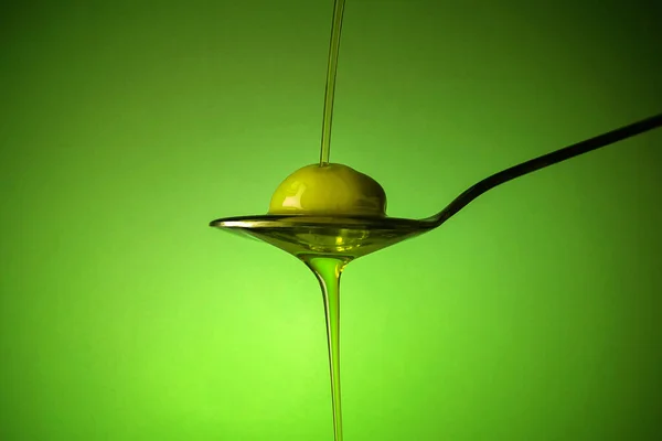 En sked med en oliv under ett fall av olja på grön bakgrund — Stockfoto