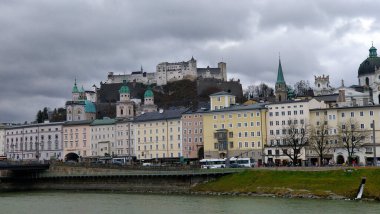 Soğuk bulutlu kış zamanı, Salzburg Şatosu 'nun ön planda nehir ve tarihi yerleşim binalarıyla güzel bir manzarası var..