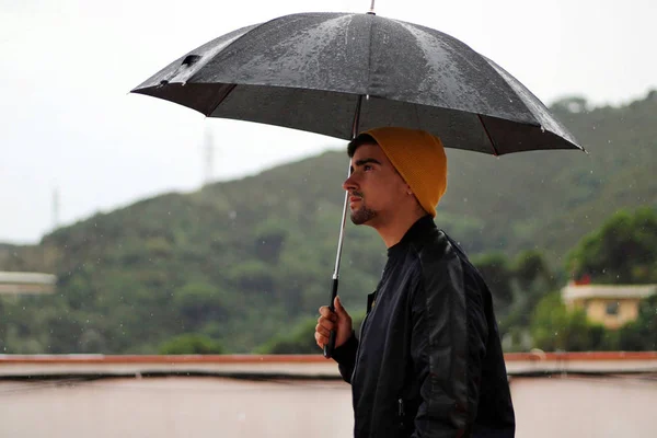 Man with a yellow wool cap and a leather jacket holds open black umbrella in his hand under the rain.
