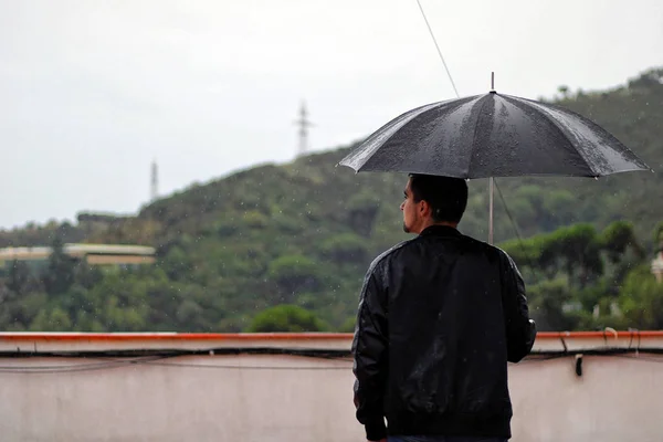 Man with a leather jacket holds open black umbrella in his hand under the rain.