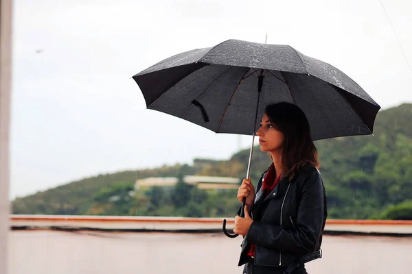 Young woman with a leather jacket holds open black umbrella in his hand under the rain.