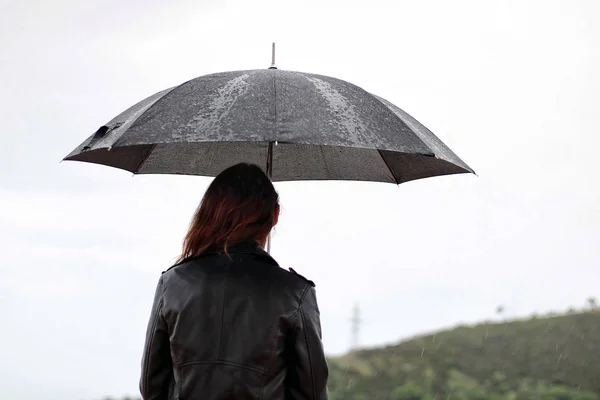 Young woman with a leather jacket holds open black umbrella in his hand under the rain.