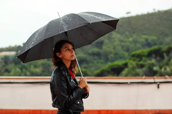 Young woman with a leather jacket holds open black umbrella in his hand under the rain.