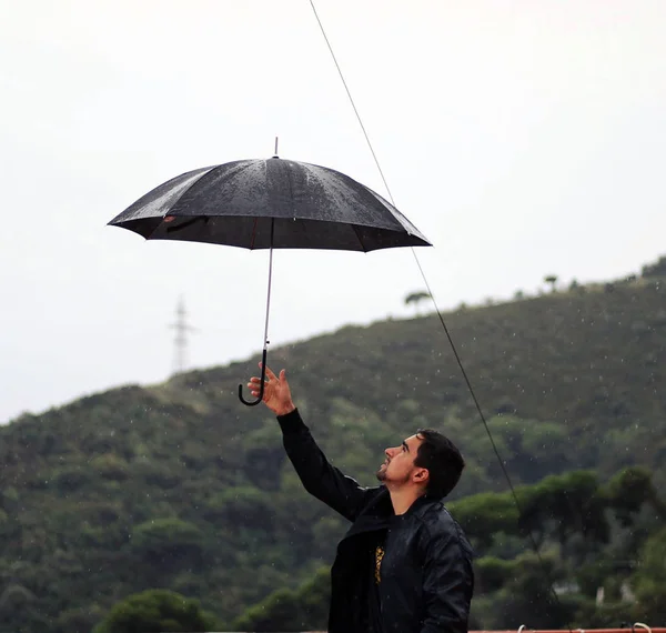 Man with a leather jacket holds open black umbrella in his hand under the rain.