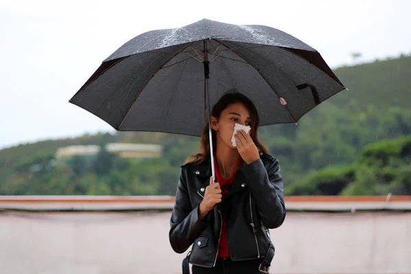 Young woman with a leather jacket holds open black umbrella in his hand under the rain and blowing the snot with a handkerchief.