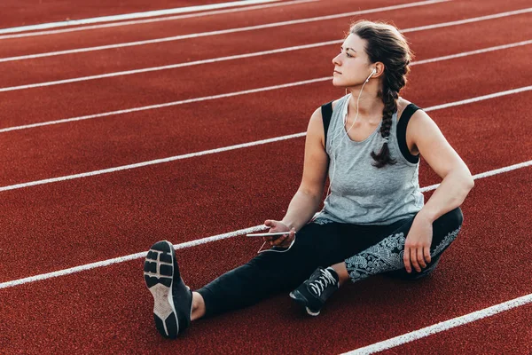 A young beautiful woman having rest with her smartphone sitting at a stadium running track.