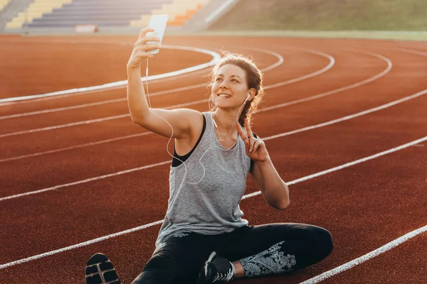A young beautiful woman making selfie sitting at a stadium running track.