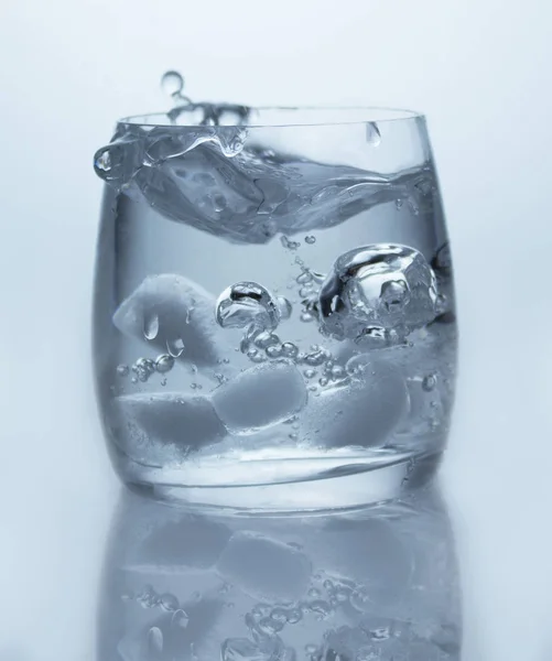 Glas met helder zuiver water, ijsblokjes, bubbels en een splash, close-up, witte achtergrond, reflectie — Stockfoto