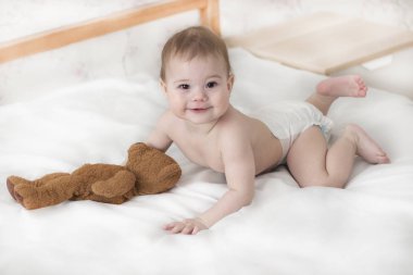 Cheerful baby boy girl in a diaper lying with a teddy bear. Cute baby in a diaper crawling on the bed, look into the camera clipart