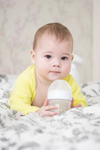 Pequeno menino menina de 7 meses segurando uma garrafa de leite. Bonito europeu branco criança em amarelo deitado na cama com uma garrafa, vertical — Fotografia de Stock